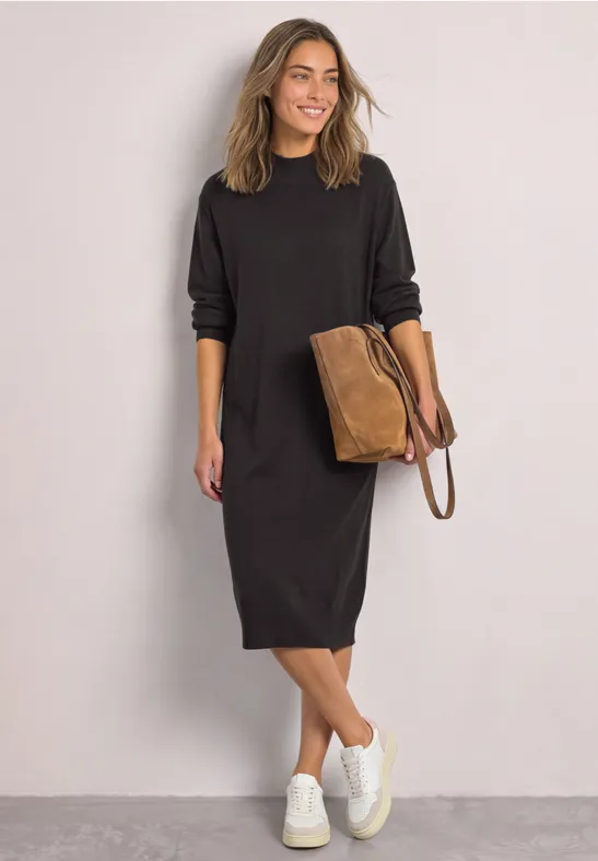 Vrouw in losse zwarte midijurk, witte sneakers en bruine tas, glimlachend voor een grijze achtergrond.