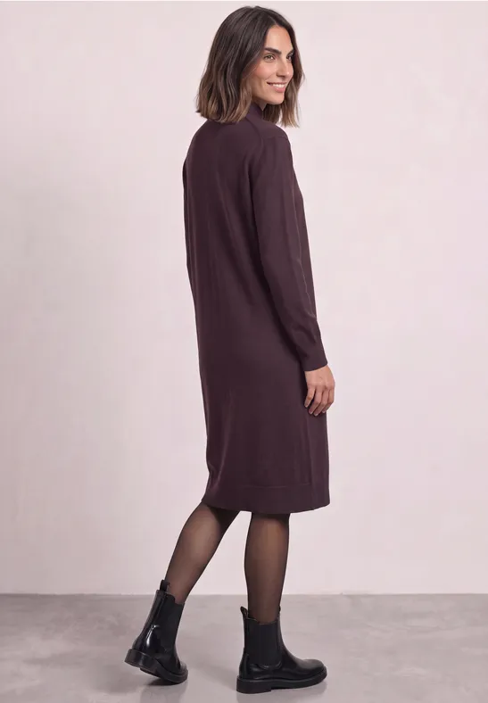 Femme en robe midi violet foncé à manches longues, collants et bottines noires, posée de profil.