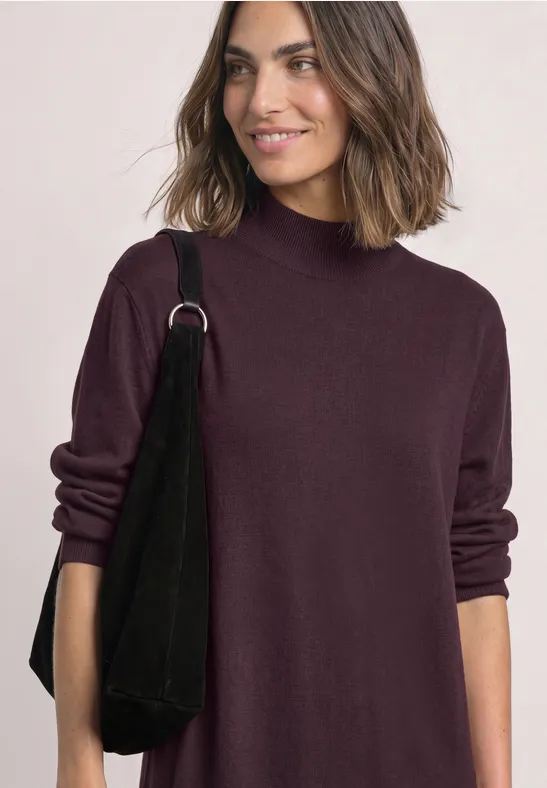 Femme souriante en pull à col roulé bordeaux, sac noir sur l’épaule.