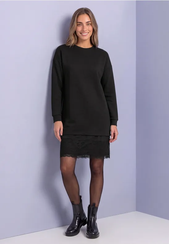 Femme en robe-pull noire à dentelle, collants noirs et bottines devant un mur violet clair.