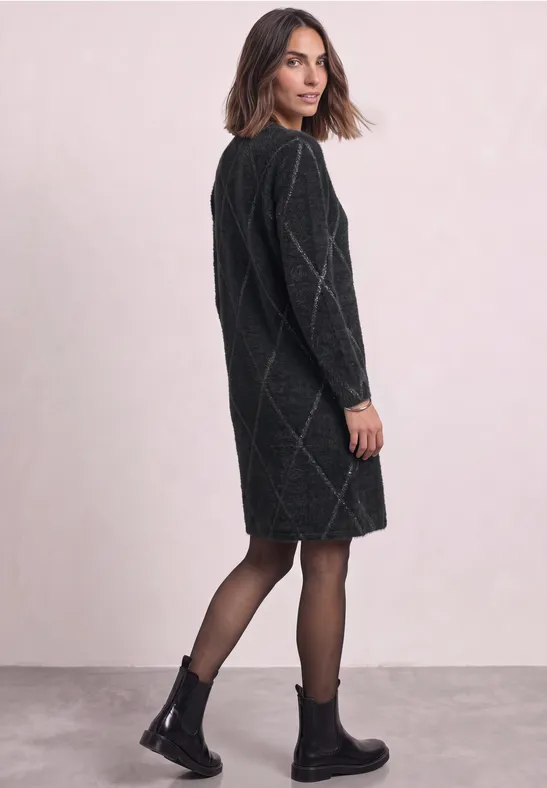 Femme en robe-pull noire à motif losange, regarde par-dessus l’épaule, porte collants et bottines noires.