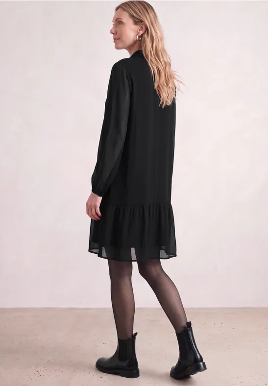 Frau in seitlicher Ansicht trägt schwarzes, lockeres Kleid mit Volant am Saum, schwarze Stiefeletten und Strumpfhose.