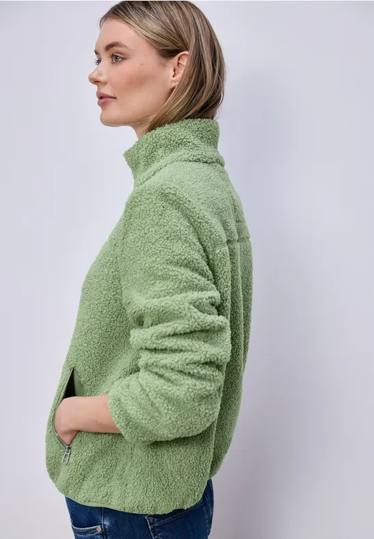 Seitliche Ansicht einer Person in grüner Fleecejacke mit Händen in den Taschen vor hellem Hintergrund.