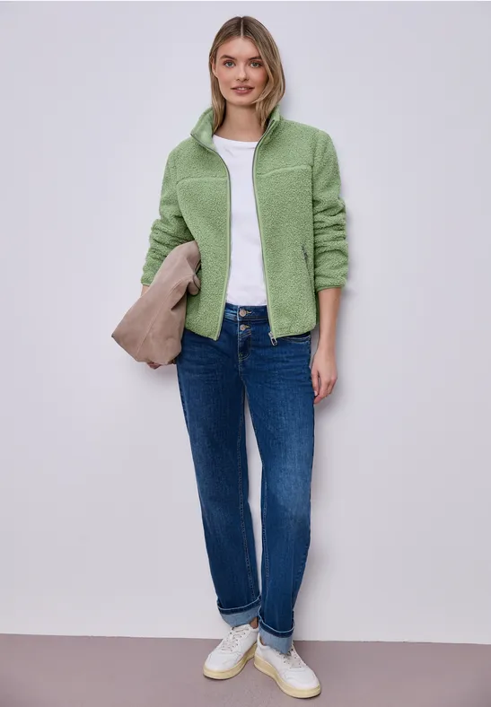 Grüne Fleecejacke, weißes T-Shirt, dunkelblaue Jeans, weiße Sneaker und beige Clutch.