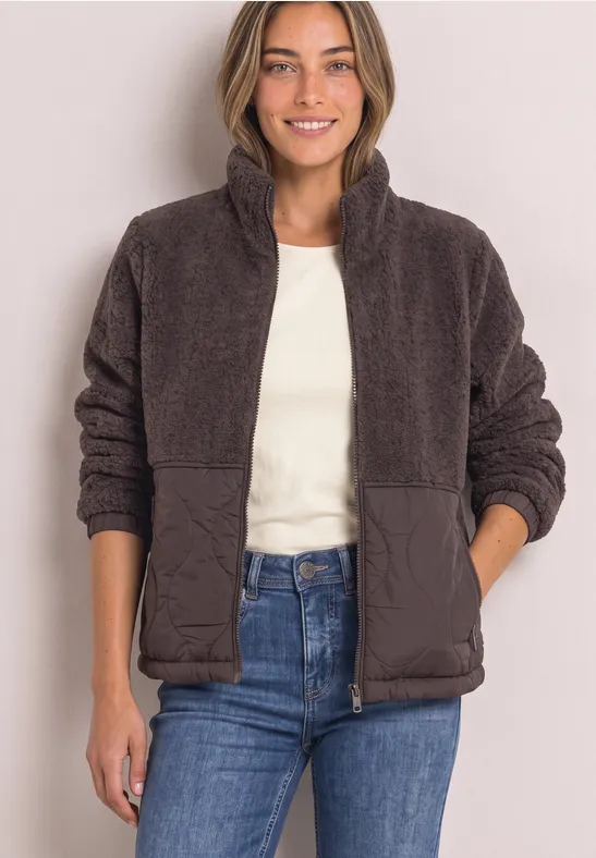 Femme souriante portant une veste en teddy marron et un jean, mains dans les poches.