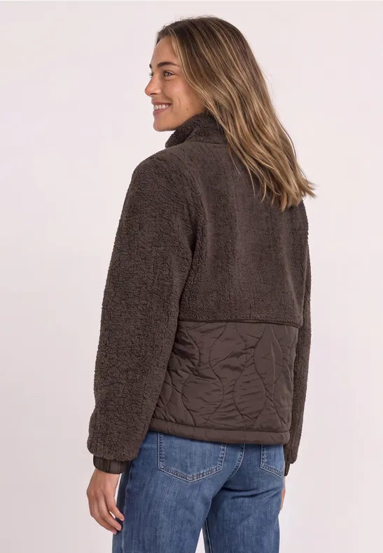Femme en veste polaire marron foncé avec dos matelassé, tourne légèrement la tête en souriant, porte un jean bleu