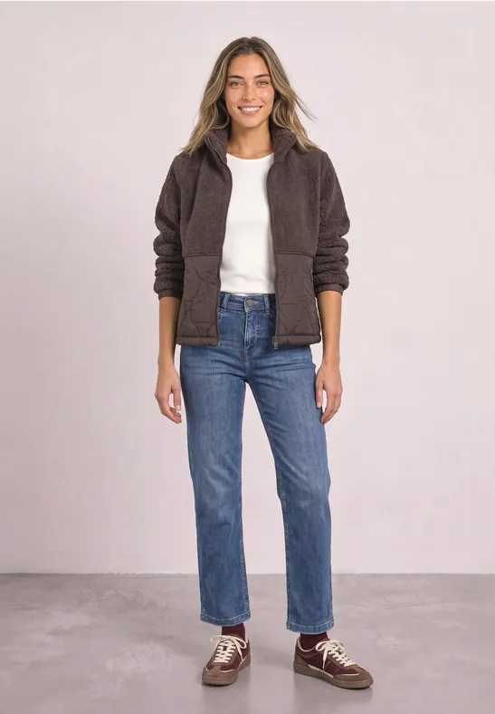 Femme souriante en jean, t-shirt blanc et veste polaire marron foncé posant devant un fond clair.
