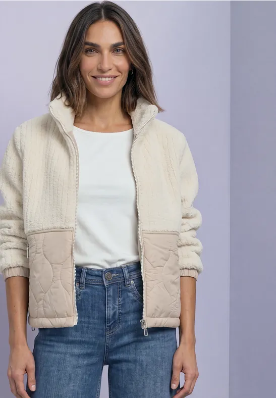 Femme portant une veste polaire blanche sur un t-shirt blanc et un jean bleu, fond neutre.
