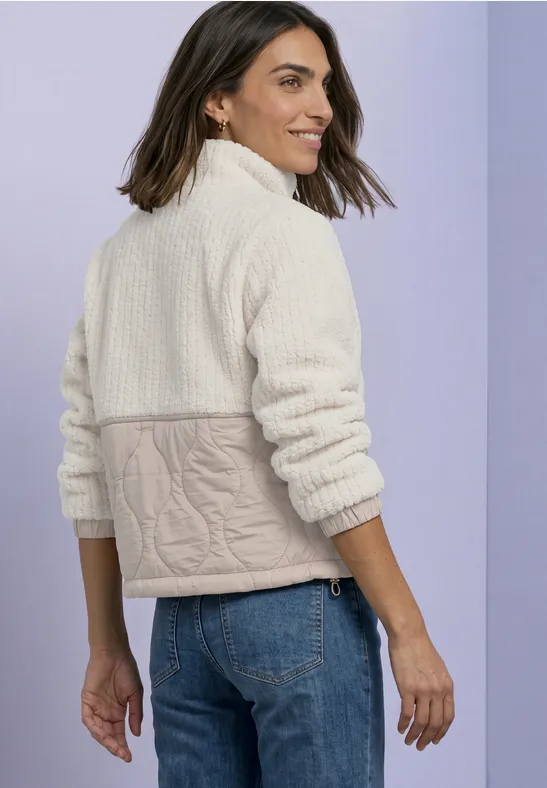 Femme en polaire crème à motif torsadé et jeans sur fond lilas, vue de profil, souriante.