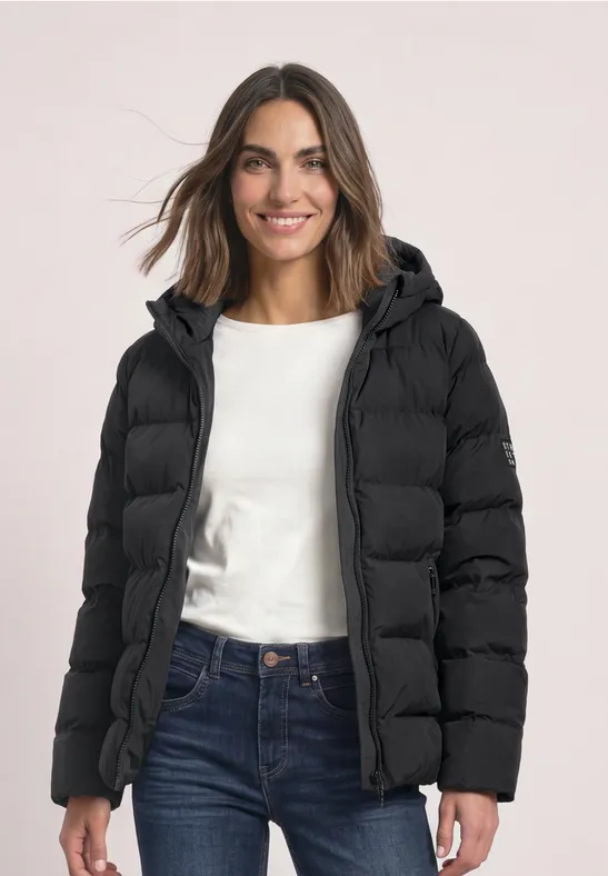 Femme souriante portant une doudoune noire à capuche, un t-shirt blanc et un jean bleu.