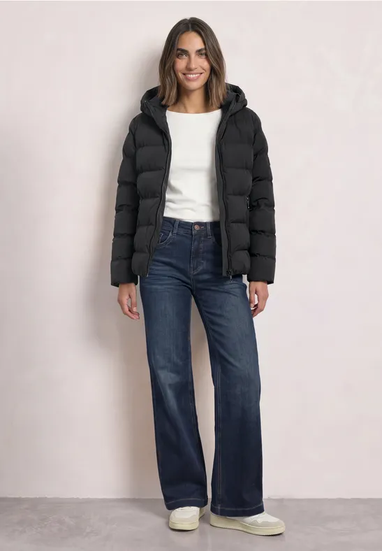 Femme souriante en doudoune noire, pull blanc et jeans larges se tenant debout.