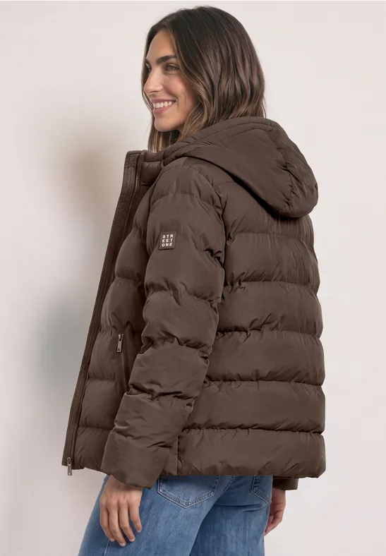 Junge Frau mit brauner Daunenjacke und Jeans, lächelnd, Blick über die Schulter vor hellem Hintergrund.