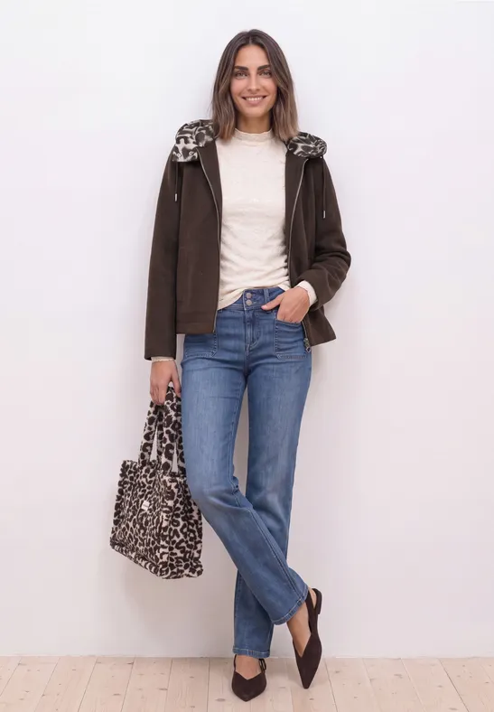 Frau in legerer Jacke, weißem Shirt, Jeans und Leopardentasche steht entspannt vor einer weißen Wand.