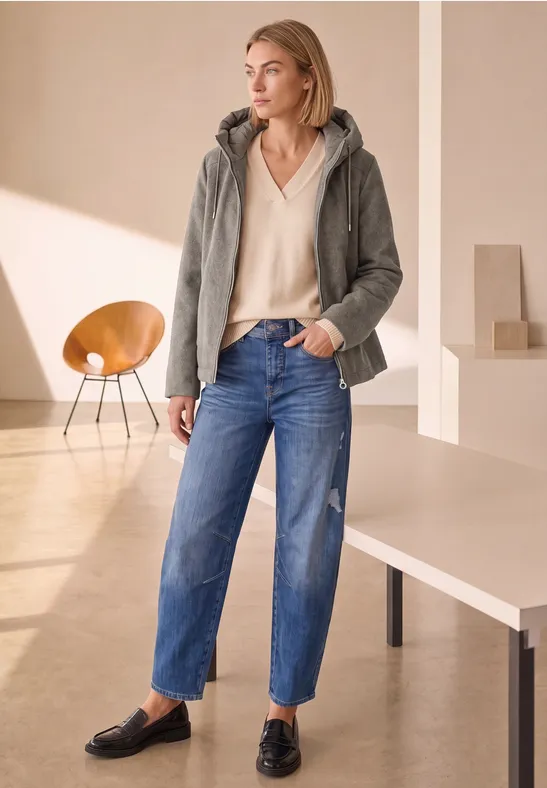 Frau in grauer Kapuzenjacke, beigem Oberteil und blauen Jeans in hellem, modernem Raum.