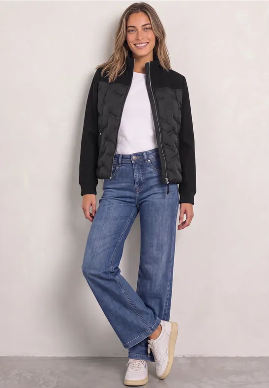 Femme souriante en veste matelassée noire, T-shirt blanc, jean bleu et baskets blanches sur fond clair.