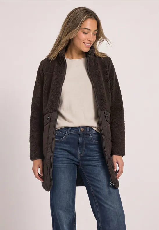 Frau in braunem Mantel, weißem Shirt und Jeans, lächelnd, blickt nach links.