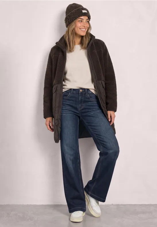 Fröhliche Frau in Beanie, weißem Shirt, braunem Mantel, weiten Jeans und weißen Sneakern vor neutralem Hintergrund.
