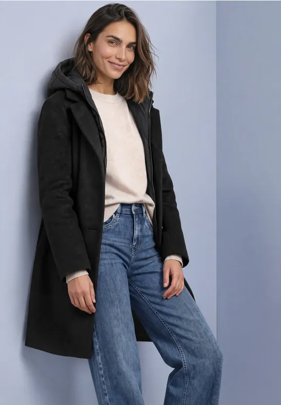 Frau in einem schwarzen Mantot, hellem Pullover und blauen Jeans, lächelnd vor neutralem Hintergrund.