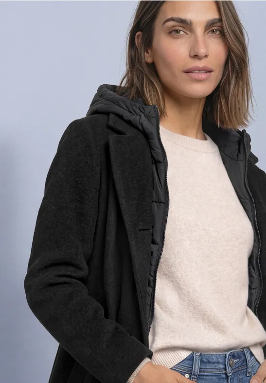 Frau mit schwarzem Kapuzenmantel, wattiertem Innenfutter, beigem Pullover und Jeans vor schlichtem Hintergrund.