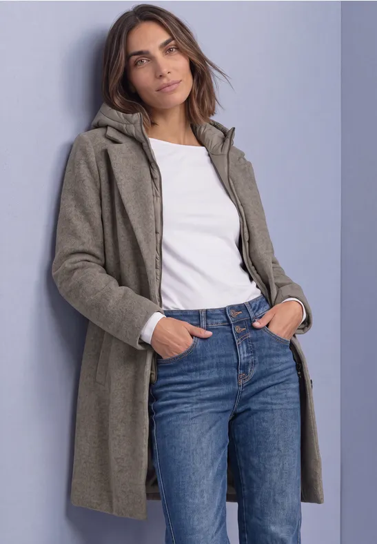 Vrouw in groengrijze jas, wit shirt en jeans, met handen in zakken tegen een muur.