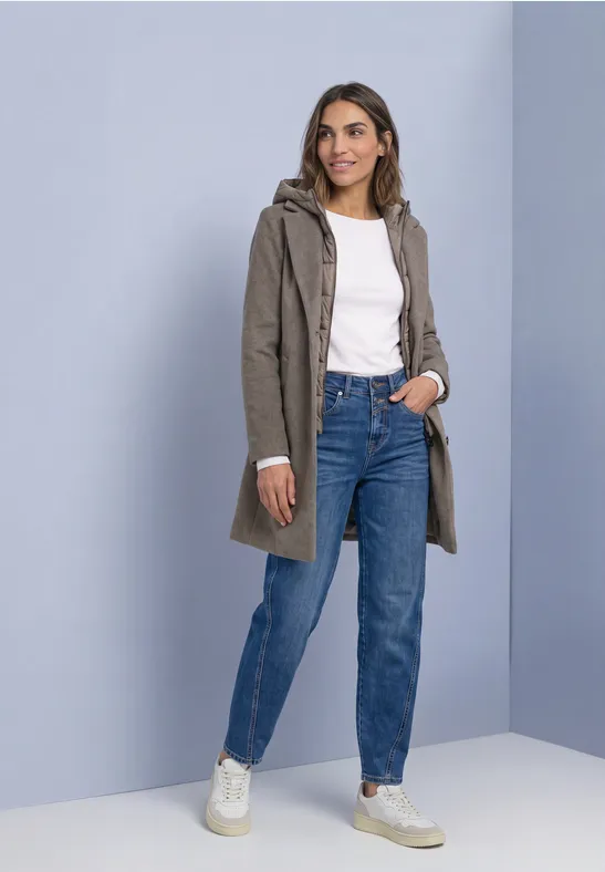 Vrouw met kaki jas met capuchon, wit shirt, blauwe jeans en witte sneakers.