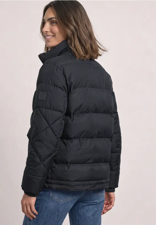 Vrouw van achteren in een zwart gewatteerd jack en blauwe jeans, glimlachend over haar schouder.