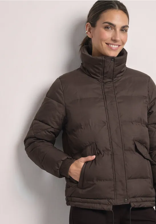 Frau trägt eine braune gesteppte Jacke mit hohem Kragen und steckt die Hände in die Taschen.