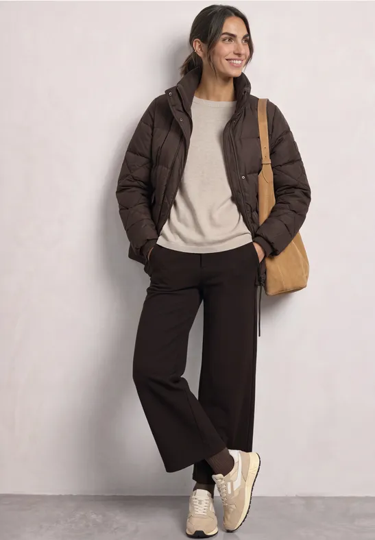 Frau lehnt an Wand, trägt braune Steppjacke, hellen Pullover, weite dunkle Hosen, Sneakers und Umhängetasche.
