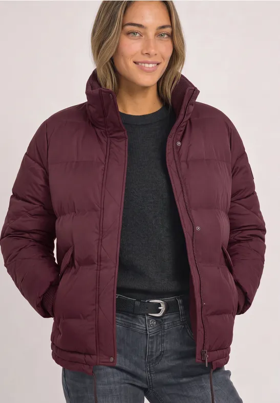 Frau in einer burgunderroten Steppjacke mit schwarzem Pullover, lächelt mit den Händen in den Taschen.