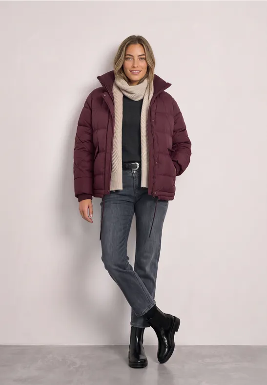 Frau in bordeauxrotem Steppjacke, grauem Schal, Jeans und Stiefeln vor hellem Hintergrund.