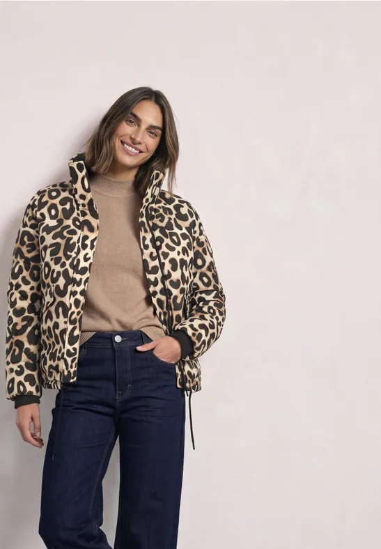 Frau in Leopardenjacke, braunem Pullover und Jeans, lächelnd vor hellem Hintergrund
