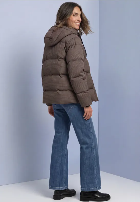 Frau in brauner Steppjacke mit Kapuze, Jeans und schwarzen Schuhen, schaut lächelnd über die Schulter.
