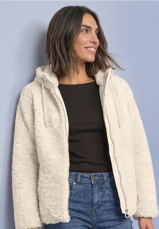 Vrouw in witte teddy-fleecejas met capuchon, zwart shirt en jeans, glimlachend en opzij kijkend.