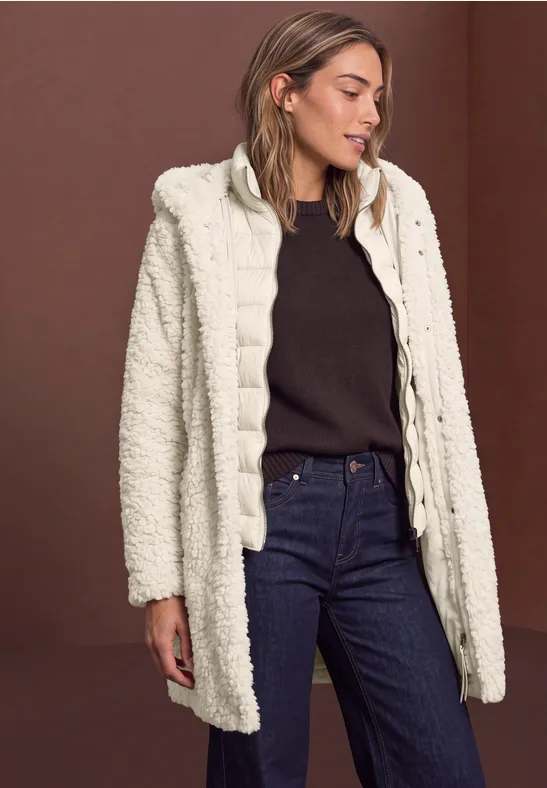 Frau trägt hellen Teddy-Mantel, dunklen Pullover und Jeans, vor braunem Hintergrund.