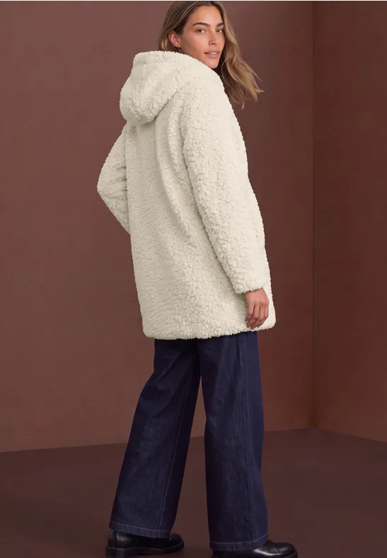 Eine Frau in weißem Teddy-Mantel mit Kapuze trägt weite Jeans und blickt seitlich vor braunem Hintergrund.