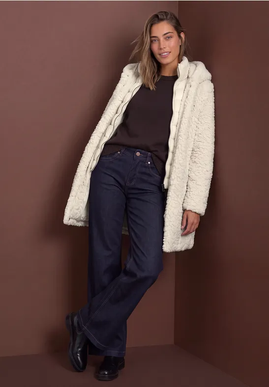 Frau in cremefarbener Teddyjacke, schwarzem Oberteil und dunkelblauen Jeans vor braunem Hintergrund.