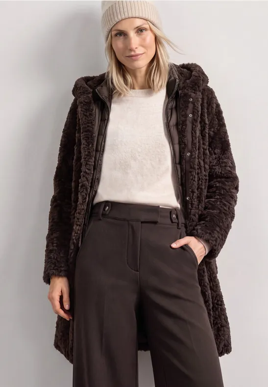 Frau mit beiger Mütze, weißem Pullover und braunem Teddy-Mantel, eine Hand in der Hosentasche.