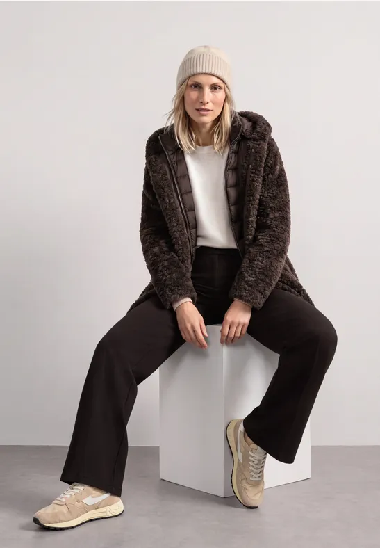 Frau mit Beanie, brauner Teddyjacke, weißem Oberteil, schwarzer Hose und hellen Sneakers sitzt lässig auf Würfel.