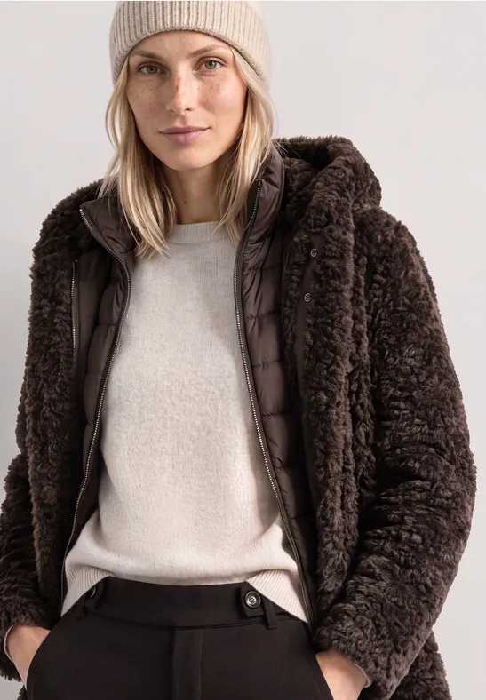 Frau mit beiger Mütze trägt braunen Teddyplüsch-Mantel mit Kapuze, hellen Pullover und dunkle Hose.