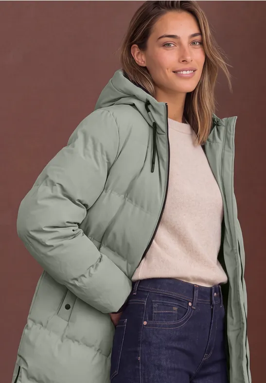 Frau in olivgrünem Steppmantel mit Kapuze, beigem Pullover und Jeans, lächelt vor braunem Hintergrund.