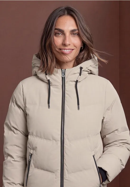 Frau mit braunen Haaren in beige-gesteppter Kapuzenjacke lächelt vor schlichtem braunem Hintergrund.