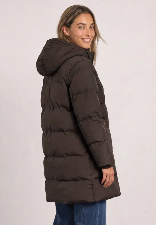 Une femme en manteau matelassé long marron à capuche, souriante, regard par-dessus l’épaule.