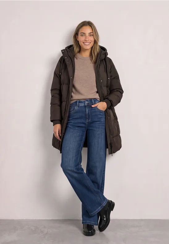 Jeune femme en manteau matelassé marron, pull beige et jean, pose souriante sur fond neutre.