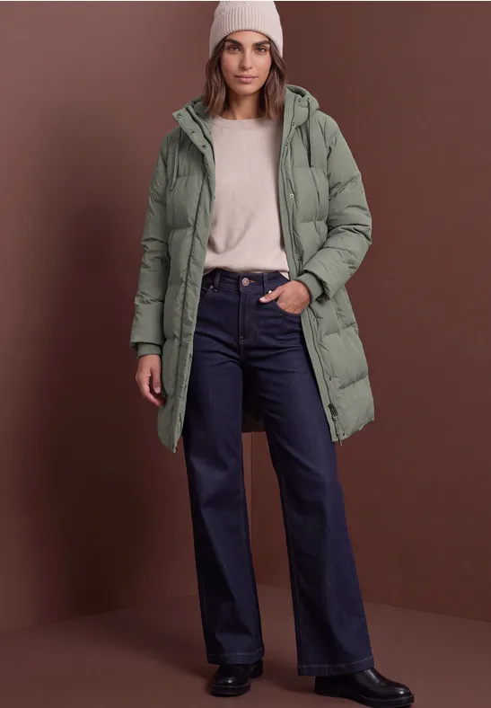 Frau in langer, grüner Steppjacke mit Kapuze, beigem Pullover, weiter Jeans und weißer Mütze stehend.