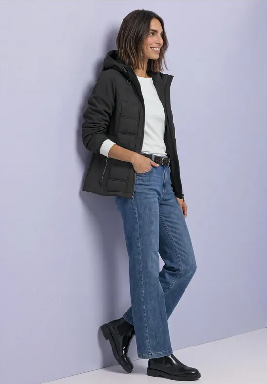 Une femme en jean et veste décontractée s'appuie sur un mur violet clair, le regard tourné de côté.