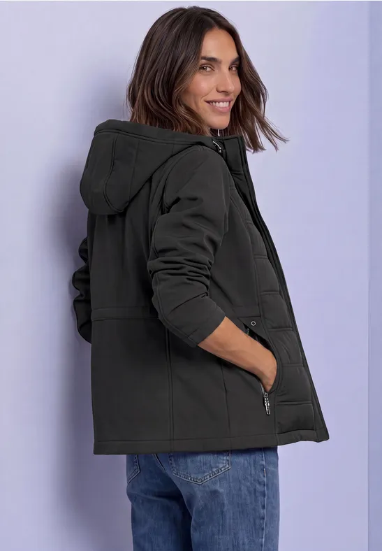 Femme en veste à capuche noire, souriante, posant de profil devant un fond clair.