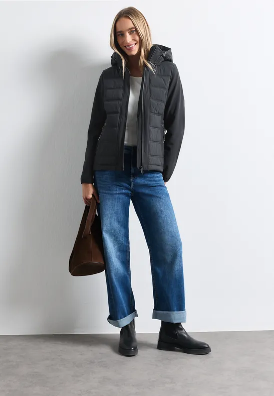 Femme en gilet matelassé noir à capuche, pull noir, jean bleu retroussé, bottines noires, sac marron.