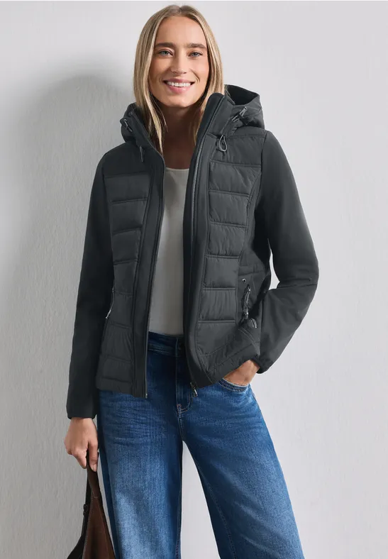 Femme souriante en veste matelassée noire à capuche, jean et sac, devant un mur clair