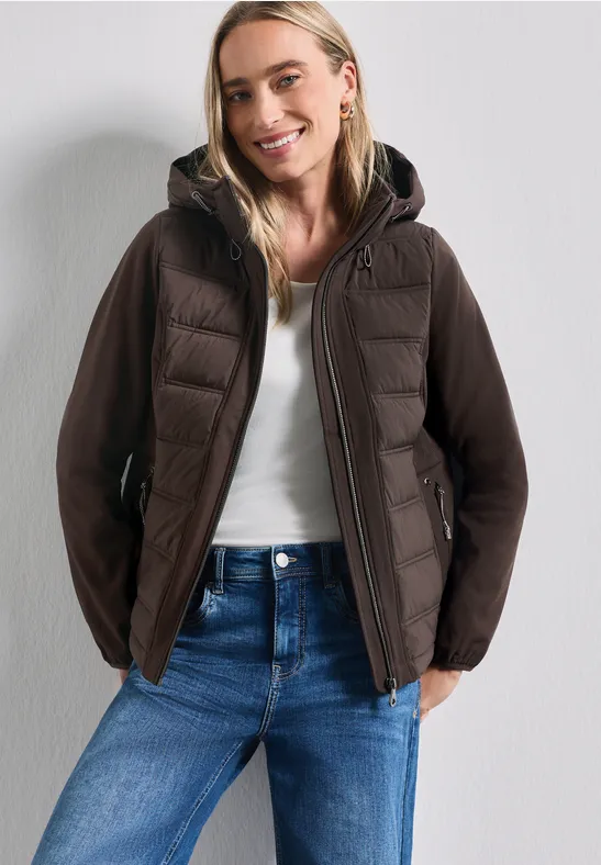 Femme souriante en top blanc, jean bleu et veste matelassée marron sur fond neutre.