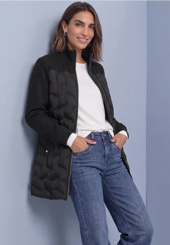 Femme en doudoune noire, top blanc et jean bleu, souriante, mains dans les poches sur fond gris.